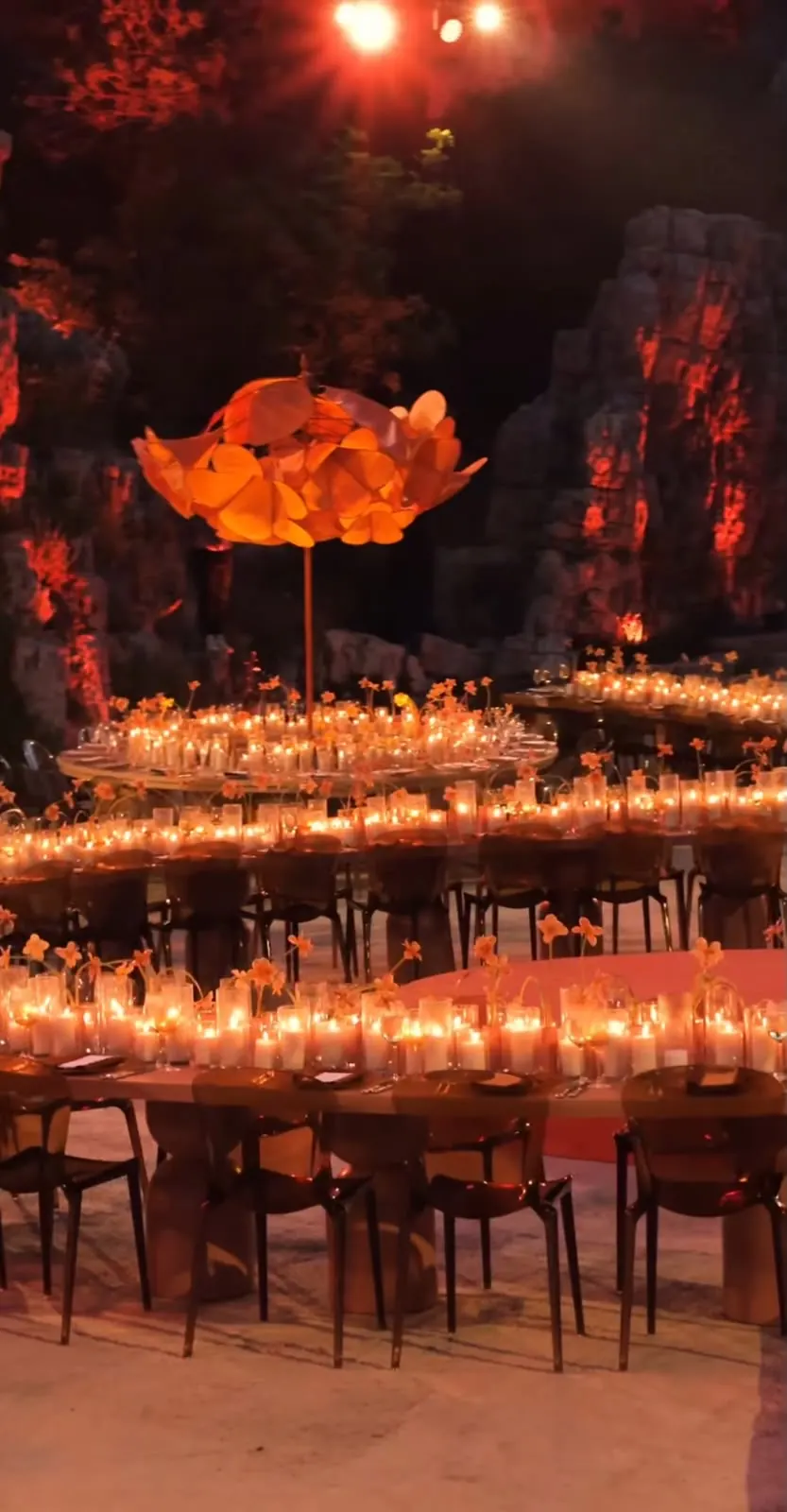 The orange flower tree glowing above rings of candlelit tables, with red-lit cliffs behind
